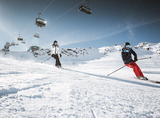 Zwei Personen fahren auf einer verschneiten Piste mit Skiliften und Bergen im Hintergrund unter einem klaren Himmel Ski.