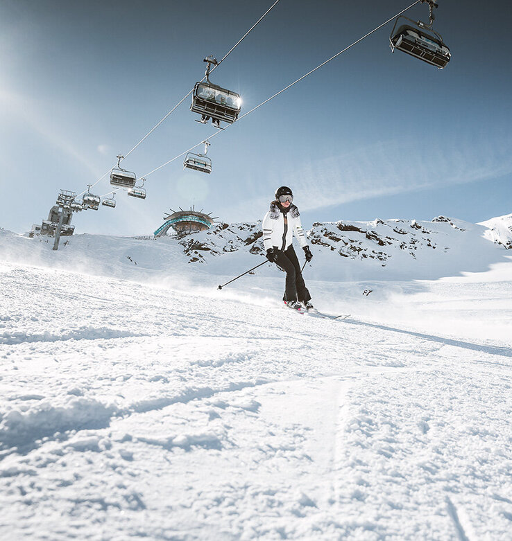Zwei Personen fahren auf einer verschneiten Piste mit Skiliften und Bergen im Hintergrund unter einem klaren Himmel Ski.