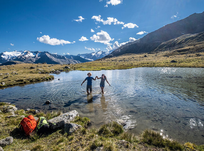 Zwei Menschen stehen händchenhaltend in einem Bergteich, im Hintergrund schneebedeckte Gipfel und blauer Himmel.