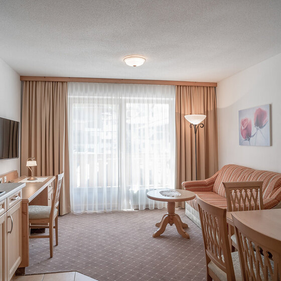 Ein heller, gemütlicher Wohnbereich mit einer Küchenzeile, einem Esstisch und Stühlen. Ein Sofa, ein Couchtisch und ein Fernseher stehen vor großen, mit beigen Vorhängen bedeckten Fenstern, die viel natürliches Licht hereinlassen. Ein Blumengemälde schmückt die Wand.