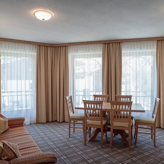 Ein heller Wohn-Essbereich mit großen, mit Vorhängen versehenen Fenstern, einem gestreiften Sofa, einem hölzernen Esstisch mit sechs Stühlen, einer Stehlampe und einem an der Wand befestigten Fernseher auf einem Teppichboden.