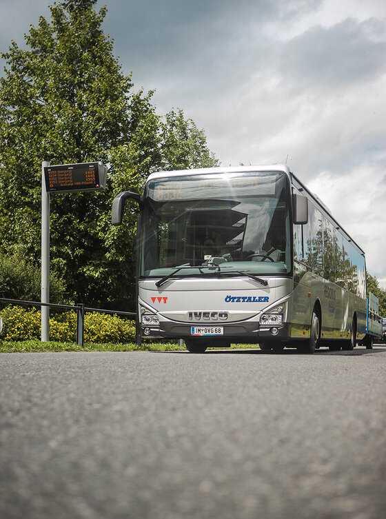 Ein silberner Reisebus parkt in der Nähe eines Bürgersteigs unter einem bewölkten Himmel, mit Bäumen und einer digitalen Anzeigetafel im Hintergrund.