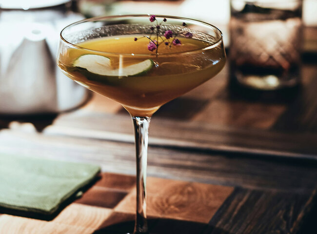 Ein Cocktail in einem Glas, garniert mit kleinen lila Blumen und einer Limettenscheibe, steht auf einem Holztisch mit einer grünen Serviette und einem verschwommenen Getränk im Hintergrund.