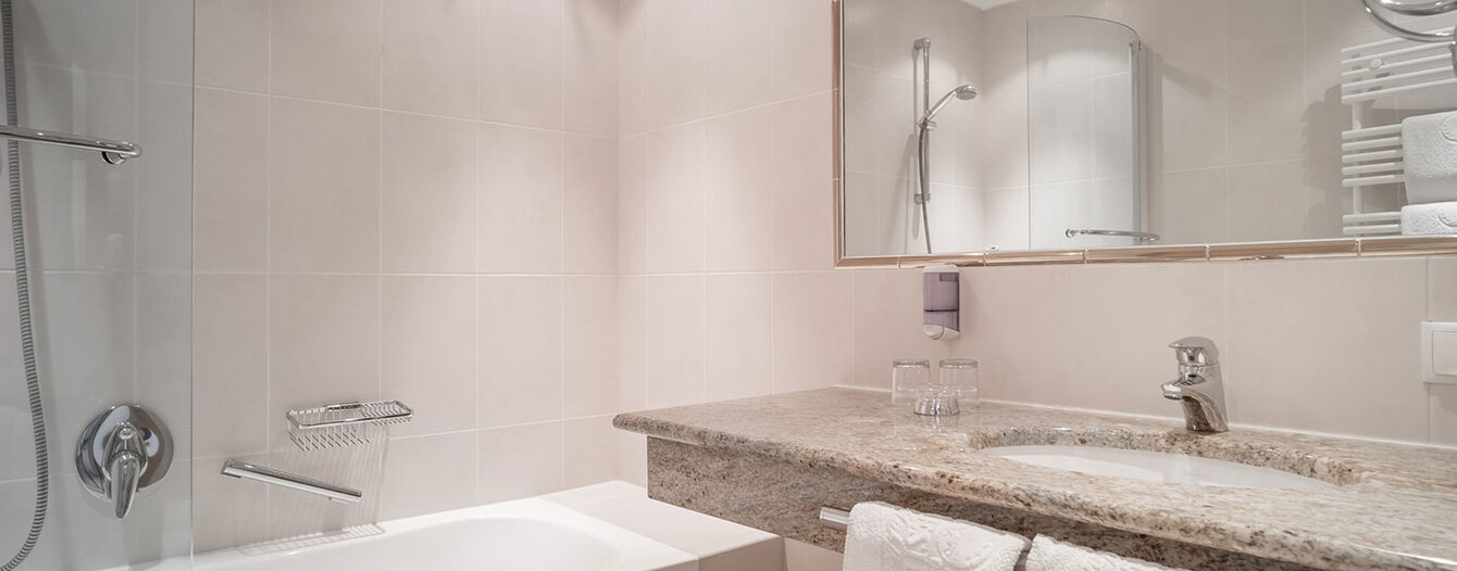 Ein modernes Badezimmer mit einer Badewanne und einer Dusche, einem großen Spiegel über einer Granitplatte mit einem Waschbecken, zwei Gläsern und ordentlich gefalteten weißen Handtüchern. Die Fließen sind in hellen Farben gehalten.