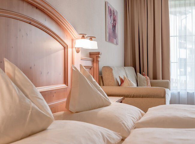 Ein gemütliches Apartmentzimmer mit einem Bett mit weißer Bettwäsche und dreieckigen Kissen, einem hölzernen Kopfteil, einer Wandlampe, einem beigen Sofa mit Kissen, einem Gemälde und großen Fenstern mit Vorhängen, die Tageslicht hereinlassen.