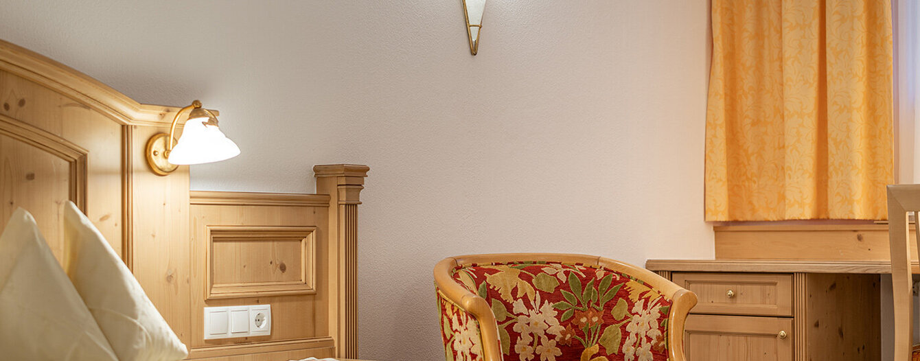 Ein gemütliches Apartmentzimmer mit einem Holzbett, einem geblümten Sessel, einem Holzschreibtisch, gelben Vorhängen und sanfter Beleuchtung durch Wandlampen.