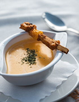 Auf einem weißen Teller steht eine Schüssel mit gelber Cremesuppe, die mit Kräutern garniert ist. Über der Schüssel erkannt man einen Spieß. Ein Löffel liegt neben der Schüssel auf einer weißen Serviette.