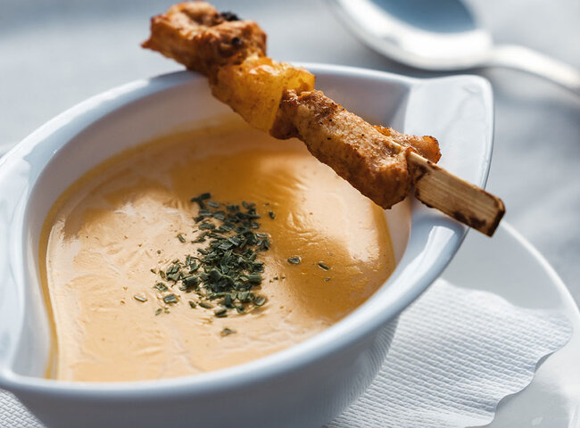 Auf einem weißen Teller steht eine Schüssel mit gelber Cremesuppe, die mit Kräutern garniert ist. Über der Schüssel erkannt man einen Spieß. Ein Löffel liegt neben der Schüssel auf einer weißen Serviette.