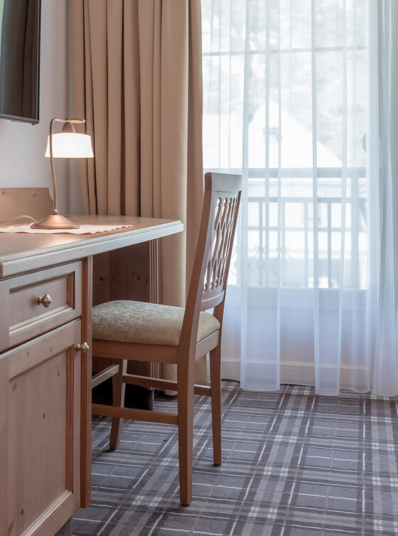 Ein gemütliches Apartmentzimmer mit einem Schreibtisch und einem Stuhl aus Holz, einer Schreibtischlampe, einem an der Wand montierten Fernseher, beigen Vorhängen und einem großen Fenster, das natürliches Licht hereinlässt. Der Boden ist mit einem gemusterten Teppich ausgelegt.