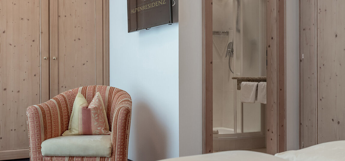 Ein gemütliches Apartmentzimmer mit einem gemusterten orangefarbenen Sessel und Kissen, einem hölzernen Kleiderschrank, einem an der Wand befestigten Fernseher und einem modernen Badezimmer mit einer durch eine offene Tür sichtbaren Glasduschkabine.