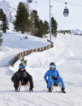 Zwei Personen mit Helmen und Winterausrüstung rodeln auf Schnee mit Skiliften und verschneiten Bergen im Hintergrund.