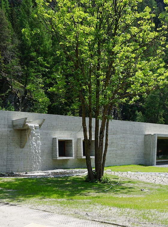 Ein modernes, minimalistisches Betongebäude mit großen Fenstern steht inmitten von saftig grünem Gras und Bäumen, umgeben von bewaldeten Hügeln bei klarem Tageslicht.