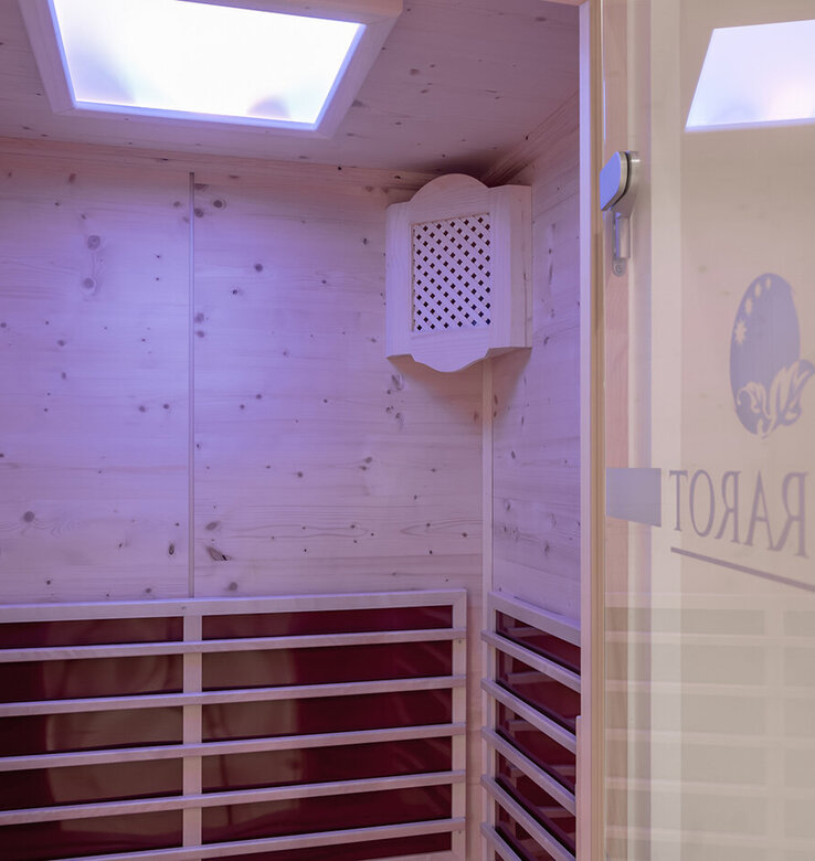 Eine hölzerne Infrarotsauna mit eingebauten Bänken, zwei belüfteten Heizpaneelen an der Wand und einem weichen, lila getönten Oberlicht über dem Kopf. Auf der Glastür ist ein Teil des Wortes INFRAROT und ein Logo zu sehen.