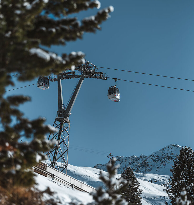 Eine Seilbahn gleitet an Seilen über eine verschneite Berglandschaft, umrahmt von Tannenbäumen mit schneebedeckten Ästen unter einem klaren blauen Himmel.