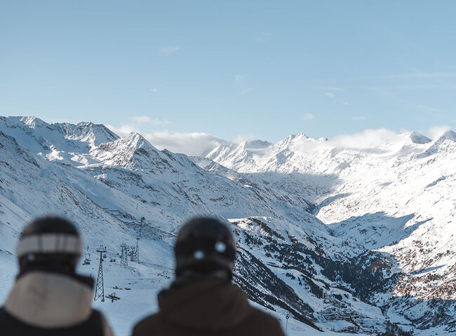 Zwei Personen in Winterjacken und Helmen stehen an einem verschneiten Berghang und blicken auf eine malerische Aussicht auf schneebedeckte Gipfel unter einem klaren blauen Himmel.