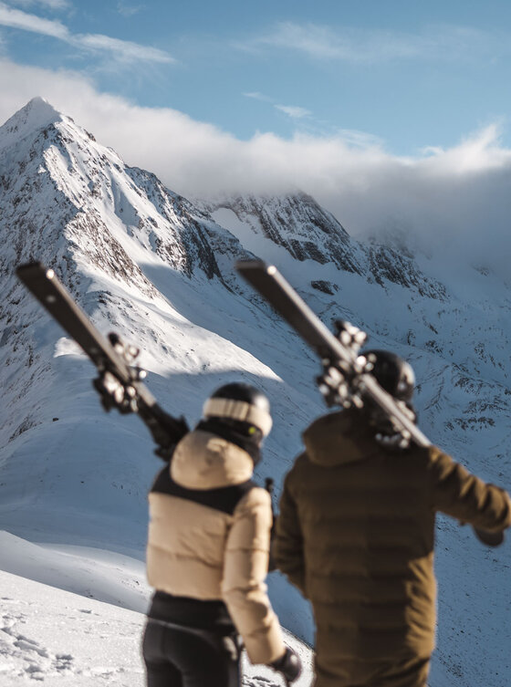Zwei Skifahrer mit Skiern blicken auf einen schneebedeckten Berg unter blauem Himmel mit vereinzelten Wolken.