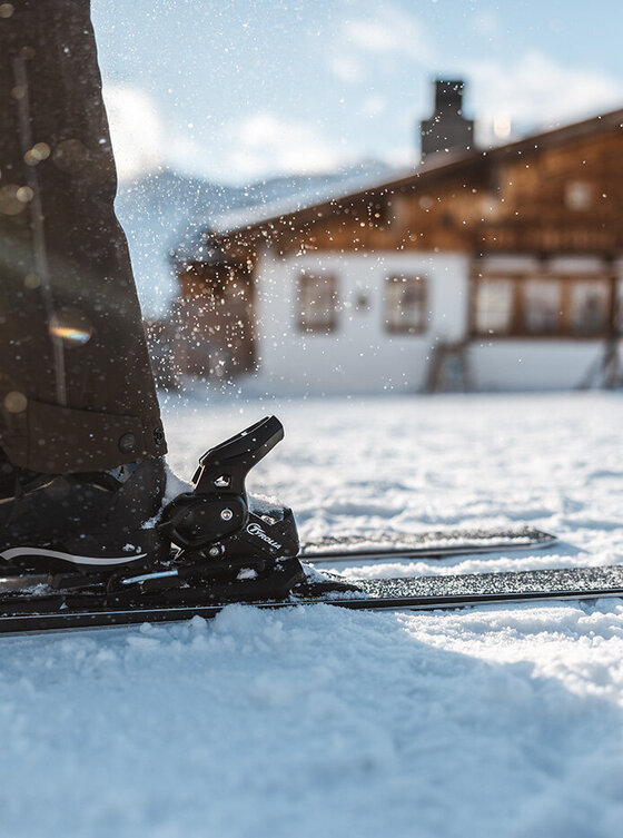 Nahaufnahme der Skischuhe einer Person auf Skiern im verschneiten Sonnenlicht, mit einer Holzhütte im Hintergrund.