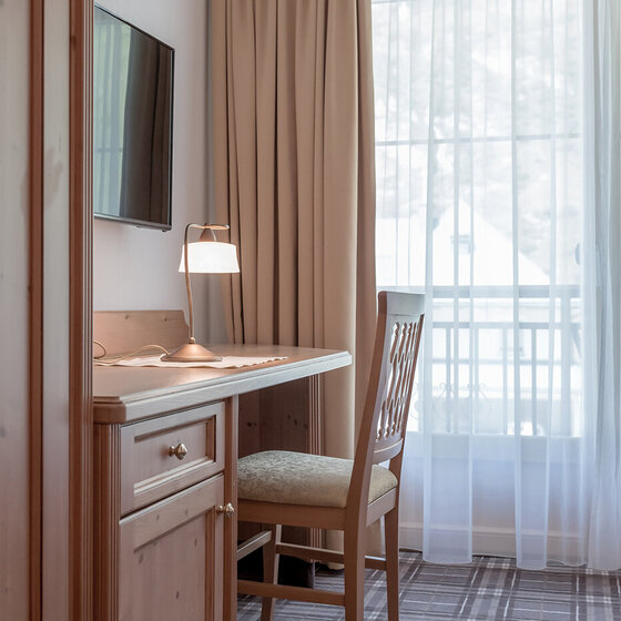 Ein hölzerner Schreibtisch mit einem Stuhl steht an einem Fenster mit weißen Gardinen und beigen Vorhängen. Eine kleine Lampe steht auf dem Schreibtisch, und an der Wand darüber ist ein Flachbildfernseher angebracht. Der Boden ist mit einem karierten Teppich ausgelegt.