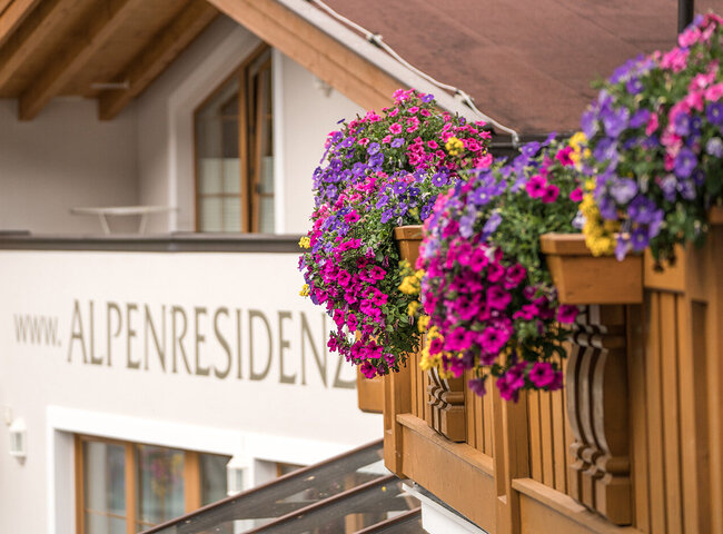 Bunte Blumenkörbe hängen an einem hölzernen Balkongeländer eines Gebäudes mit der Aufschrift "ALPENRESIDENZ" an der Wand. Das Gebäude hat weiße Wände, Holzverkleidungen und große Fenster, die an einen alpinen Stil erinnern.