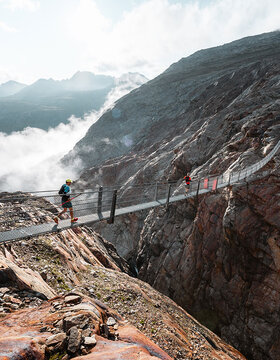 Zwei Personen mit Helmen und Wanderausrüstung überqueren eine lange Hängebrücke über eine tiefe Felsenschlucht, mit Bergen und Wolken im Hintergrund.