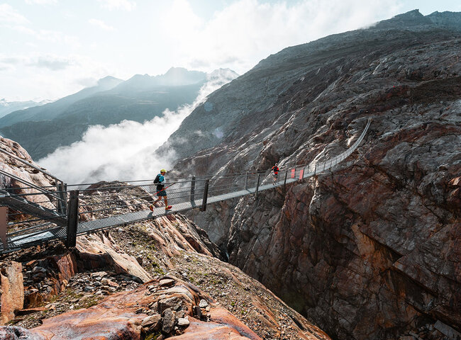 Zwei Personen mit Helmen und Wanderausrüstung überqueren eine lange Hängebrücke über eine tiefe Felsenschlucht, mit Bergen und Wolken im Hintergrund.
