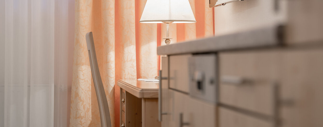 Ein sanft beleuchteter Raum mit einem Schreibtisch und einem Stuhl, einer eingeschalteten Tischlampe, hellen Vorhängen und einem Schrank im Vordergrund. Warme und gemütliche Atmosphäre.