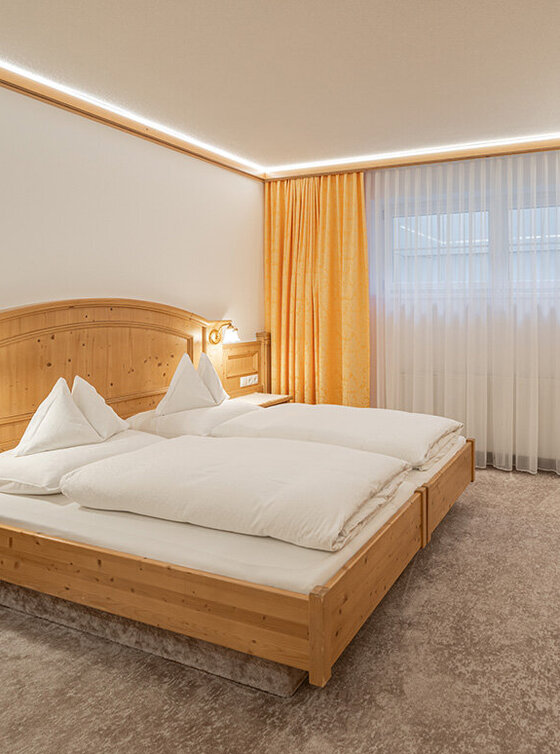 Ein gemütliches Apartmentzimmer mit einem Doppelbett, Holzmöbeln, einem Schreibtisch mit Lampe, einem an der Wand montierten Fernseher und einer Küchenzeile mit Kaffeemaschine. Ein großes Fenster mit durchsichtigen und gelben Vorhängen lässt natürliches Licht herein.