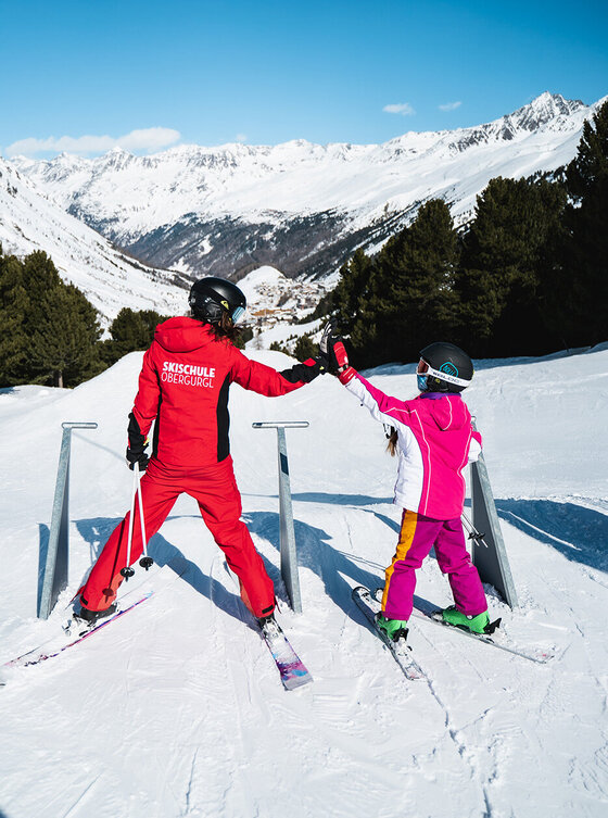 Skilehrer und Kind stehen auf einem verschneiten Berghang, umgeben von Bäumen und schneebedeckten Gipfeln. Sie geben sich ein High-Five.