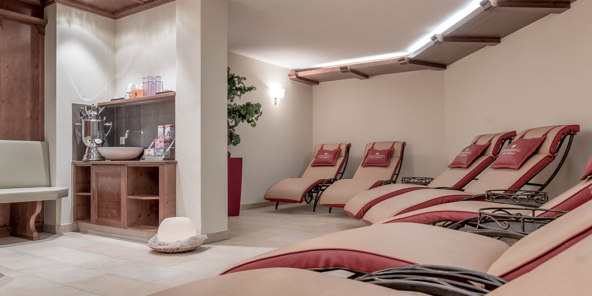 Ein ruhiger Wellness-Raum mit mehreren gepolsterten Liegestühlen, einem kleinen hölzernen Waschbecken mit Handtüchern und Wasser und sanfter Beleuchtung. Die Atmosphäre ist ruhig und einladend.