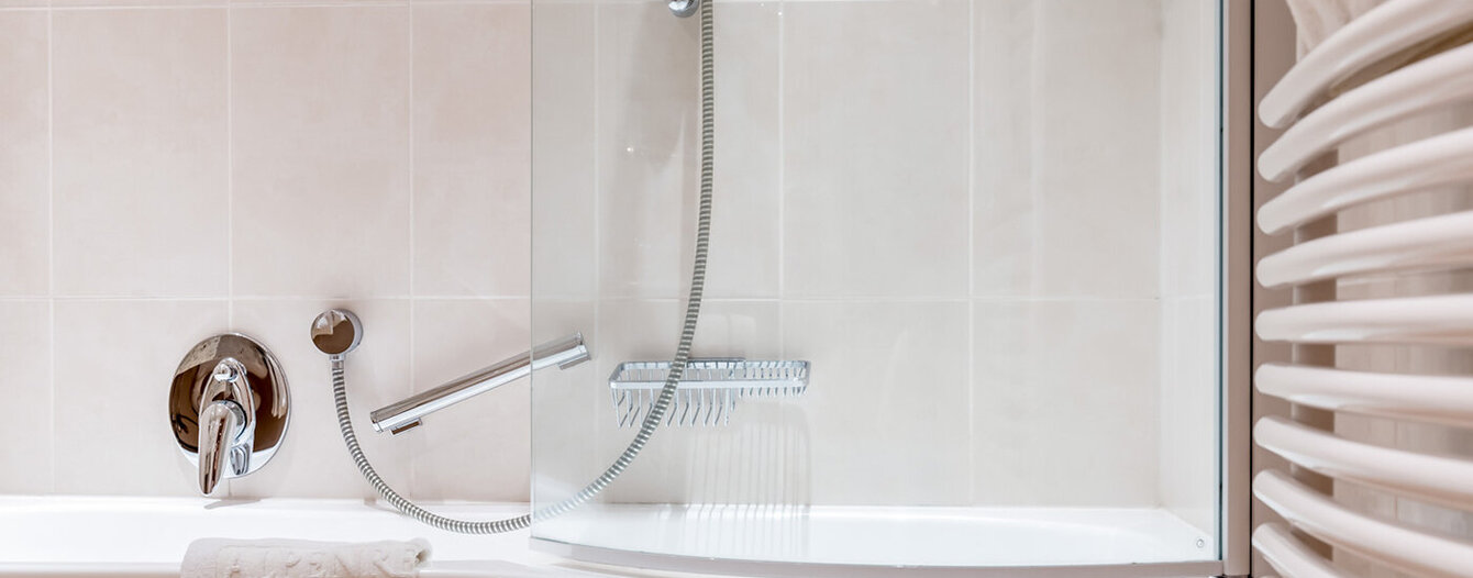 Modernes Badezimmer mit weißer Badewanne, Duschkopf, Glasduschwand, Handtuchhalter und einem gefalteten weißen Handtuch, das über dem Wannenrand hängt. Die Wände und Fliesen sind hellbeige.