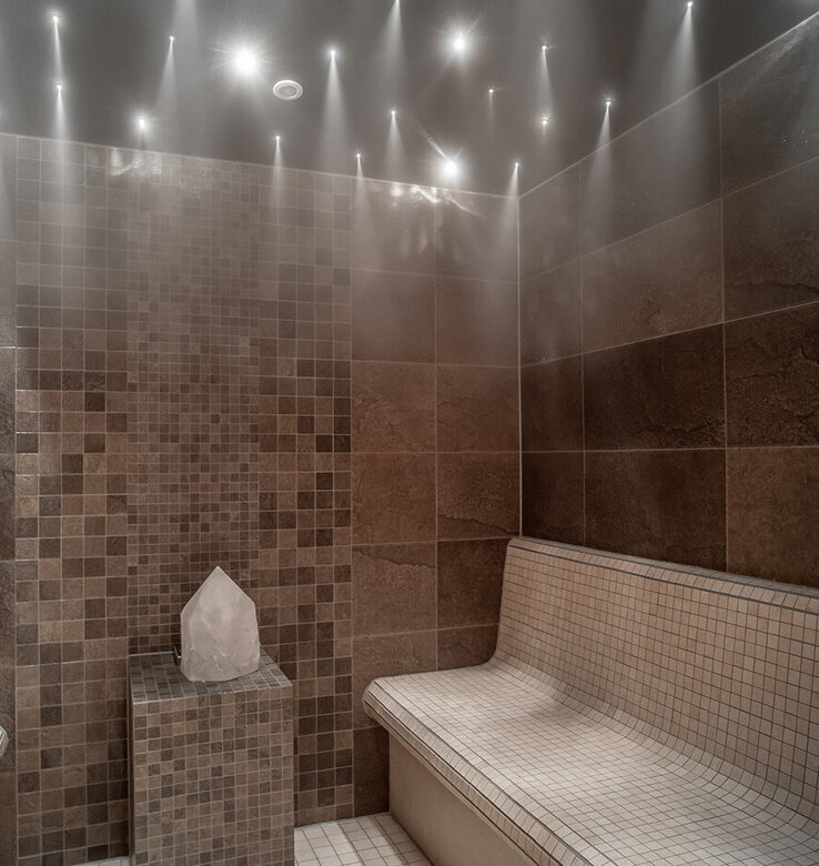 Ein modernes Dampfbad mit zwei gefliesten Bänken, braunen und beigen Mosaikwänden, einem Kristall auf einem Sockel und einer Deckenbeleuchtung, die sanfte Strahlen durch den Dampf sendet.