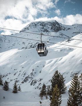 Eine Seilbahn, die an einem Seil aufgehängt ist, fährt über verschneite Berghänge, unter denen sich Kiefern befinden, mit einem schroffen, schneebedeckten Gipfel im Hintergrund und einem teilweise bewölkten Himmel.