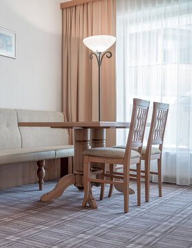 Ein heller, gemütlicher Raum mit einem runden Holztisch, zwei Holzstühlen, einer gepolsterten Bank, einer Stehlampe und großen Fenstern, die von weißen Vorhängen verdeckt werden.