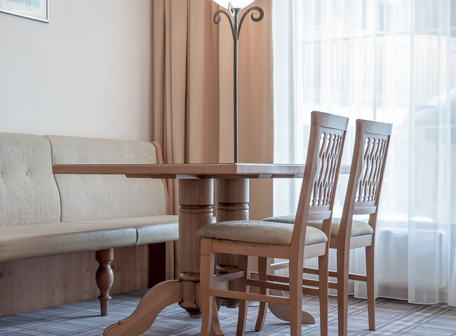 Ein heller, gemütlicher Raum mit einem runden Holztisch, zwei Holzstühlen, einer gepolsterten Bank, einer Stehlampe und großen Fenstern, die von weißen Vorhängen verdeckt werden.