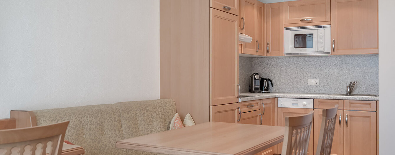Eine kleine Küche mit hellen Holzschränken, einer eingebauten Mikrowelle und einer Spüle. Davor befindet sich ein Essbereich mit einem Holztisch, vier Stühlen und einer gepolsterten Bank an der Wand. Der Raum wirkt sauber und minimalistisch.