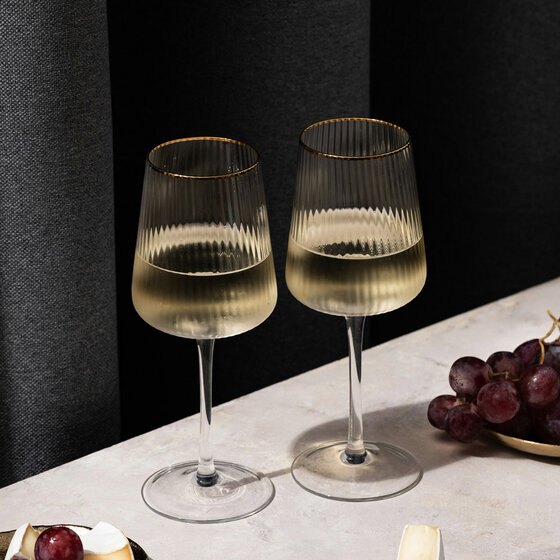 Zwei Gläser Weißwein auf einer hellen Fläche, dazu ein kleiner Teller mit Käse und roten Weintrauben, im Hintergrund ein dunkler Vorhang.