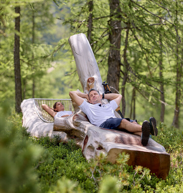Zwei Personen entspannen sich auf einer großen Holzbank in einem Wald, umgeben von grünen Bäumen und Sträuchern.