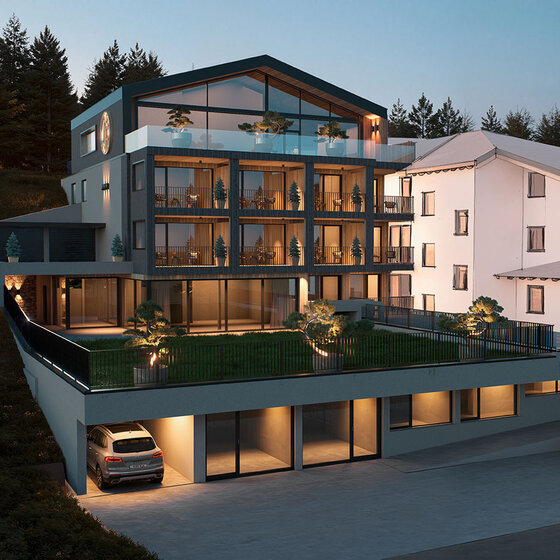 Modernes mehrstöckiges Gebäude in der Abenddämmerung mit großen Glasfenstern, Balkonen und begrünten Terrassen; zwei Autos sind in der Einfahrt geparkt, und im Hintergrund stehen Bäume. Warme Innenbeleuchtung erhellt die Szene.