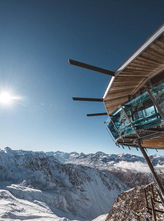 Modernes Glasgebäude auf einem schneebedeckten Berg, die Sonne scheint über die entfernten schneebedeckten Gipfel unter einem klaren Himmel.