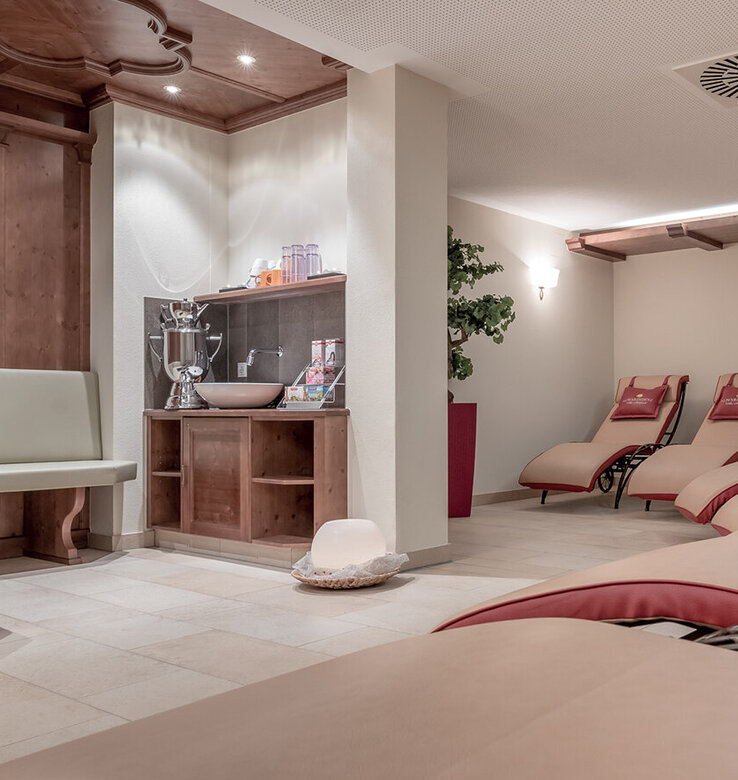 Ein ruhiger Spa-Entspannungsraum mit mehreren gepolsterten Liegen, roten Akzentkissen, sanfter Beleuchtung, einer hölzernen Teebar mit Wasser und Gläsern.