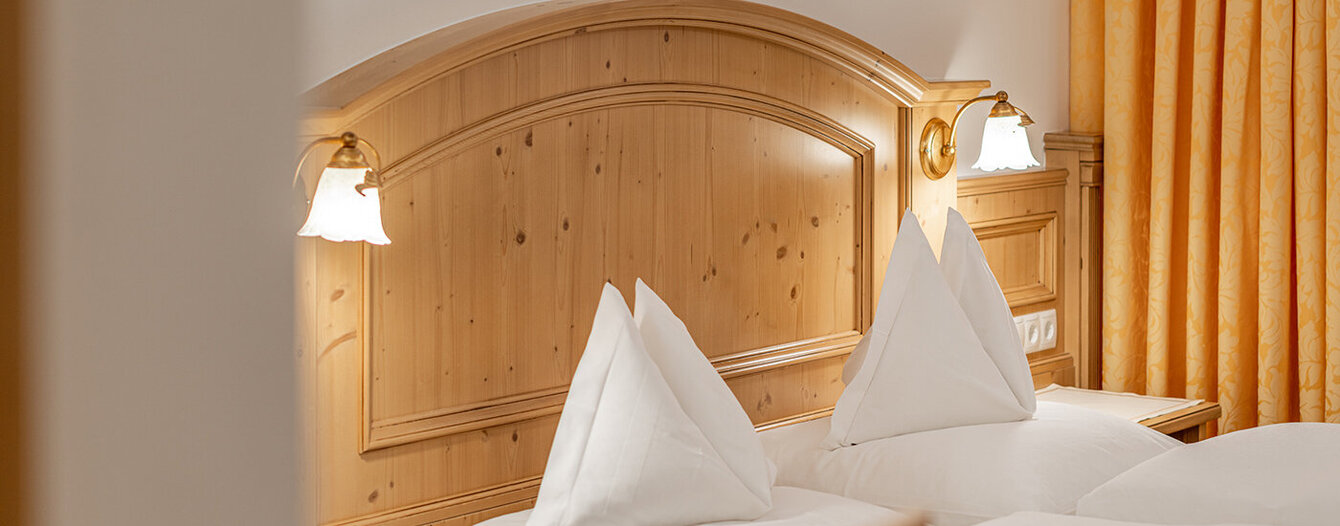 Ein aufgeräumtes Apartmentzimmer mit einem Doppelbett, weißer Bettwäsche, einem hölzernen Kopfteil, Nachttischlampen und gelb gemusterten Vorhängen.