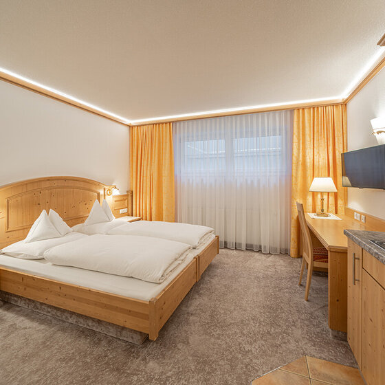 Ein gemütliches Apartmentzimmer mit einem Doppelbett, Holzmöbeln, einem Schreibtisch mit Lampe, einem an der Wand montierten Fernseher, einer Kochnische und einem großen Fenster mit gelben Vorhängen, das natürliches Licht hereinlässt.