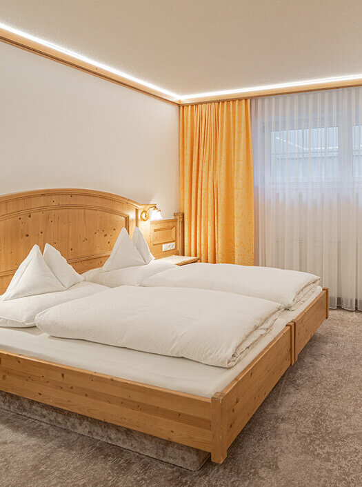 Ein gemütliches Apartmentzimmer mit einem Doppelbett, Holzmöbeln, einem Schreibtisch mit Lampe, einem an der Wand montierten Fernseher, einer Kochnische und einem großen Fenster mit gelben Vorhängen, das natürliches Licht hereinlässt.