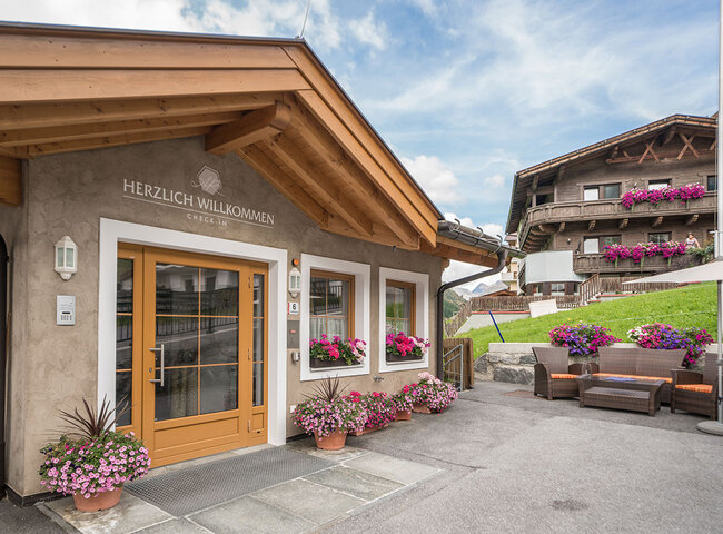 Eingang eines gemütlichen Alpengasthofs mit Blumenkästen, einer Holztür und einem Schild mit der Aufschrift Herzlich Willkommen. Im Hintergrund sind Sitzgelegenheiten im Freien und ein weiteres Haus mit Balkonen und Blumen zu sehen.