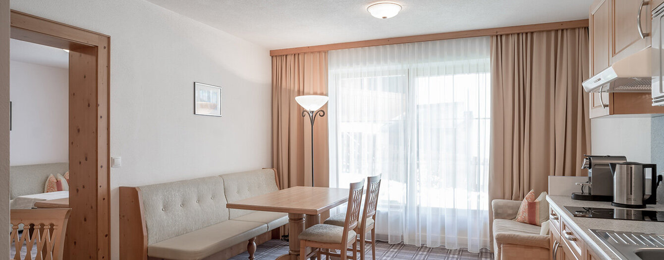 Eine helle, moderne Küche und ein Essbereich mit hellen Holzmöbeln, einer eingebauten Eckbank, einem rechteckigen Tisch, zwei Stühlen, einem Sofa und großen Fenstern mit transparenten Vorhängen. Ein Schlafzimmer ist durch eine offene Tür sichtbar.