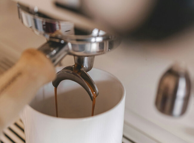 Nahaufnahme einer Espressomaschine, die frischen Espresso in eine weiße Keramiktasse einschenkt, wobei aus dem Siebträgerauslauf satte braune Kaffeeströme fließen.