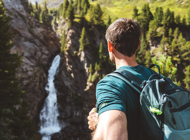 Ein Mann mit Rucksack steht an einem hellen, sonnigen Tag vor einem Wasserfall und üppigen Bergen.