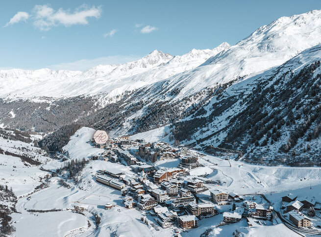 Luftaufnahme eines schneebedeckten Alpendorfs in einem Tal, umgeben von bewaldeten Hügeln und hohen, zerklüfteten Bergen unter einem klaren blauen Himmel.