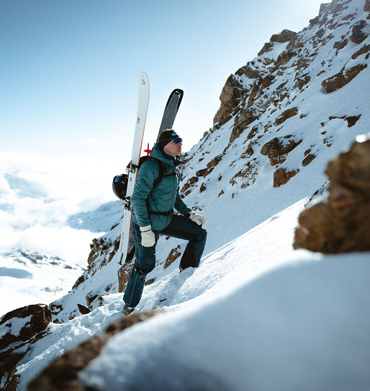 Eine Person in Winterkleidung und mit Sonnenbrille klettert einen verschneiten Berghang hinauf, wobei sie Skier auf der Schulter trägt, mit Felsen und blauem Himmel im Hintergrund.