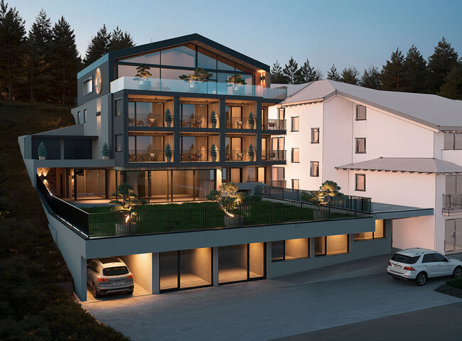 Modernes mehrstöckiges Gebäude mit großen Glasfenstern und Balkonen, in der Dämmerung warm beleuchtet. Autos sind in der Einfahrt und in der Garage geparkt. Bewaldete Hügel im Hintergrund.